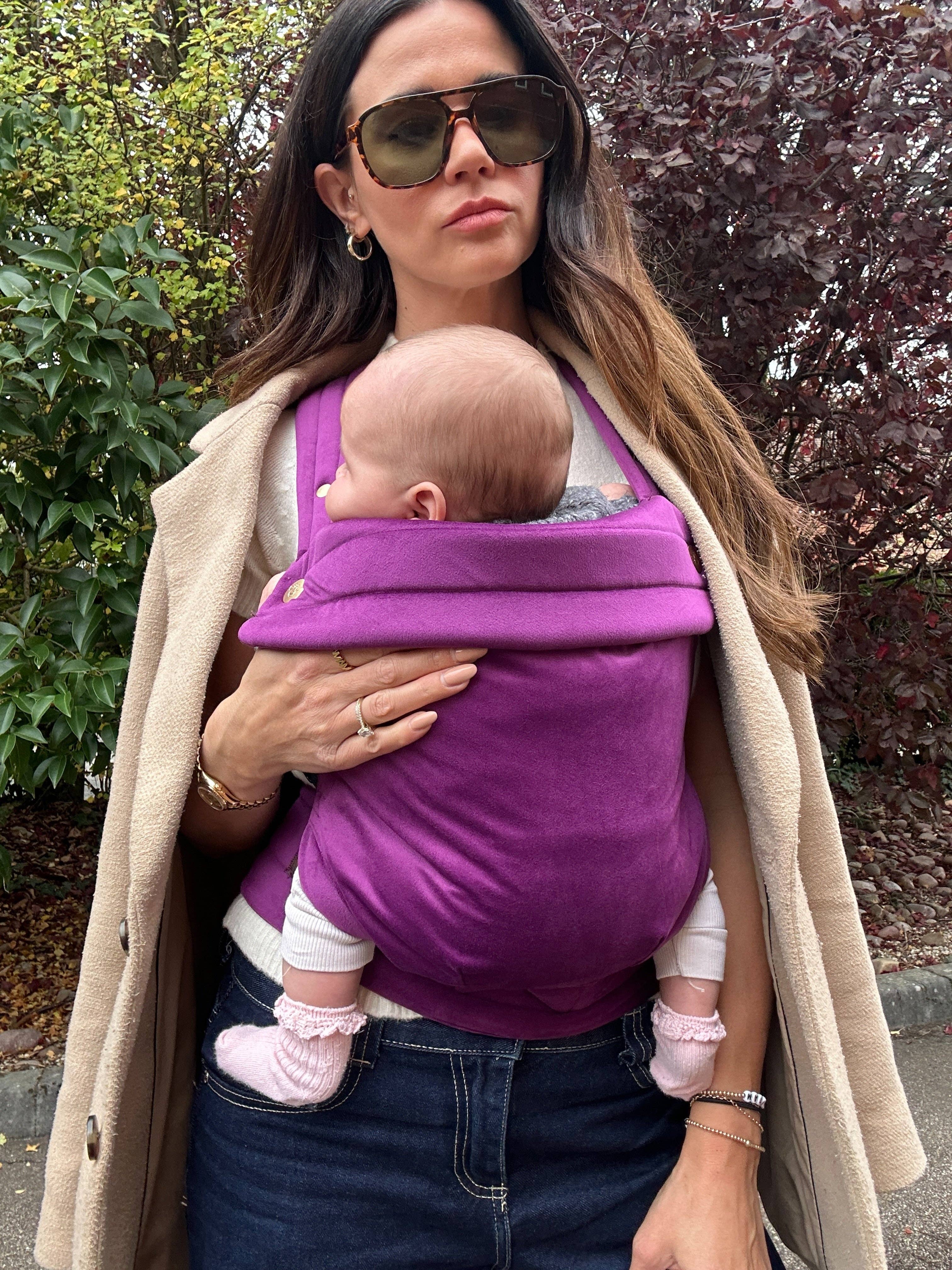 Woman holding a baby in a purple carrier outdoors.