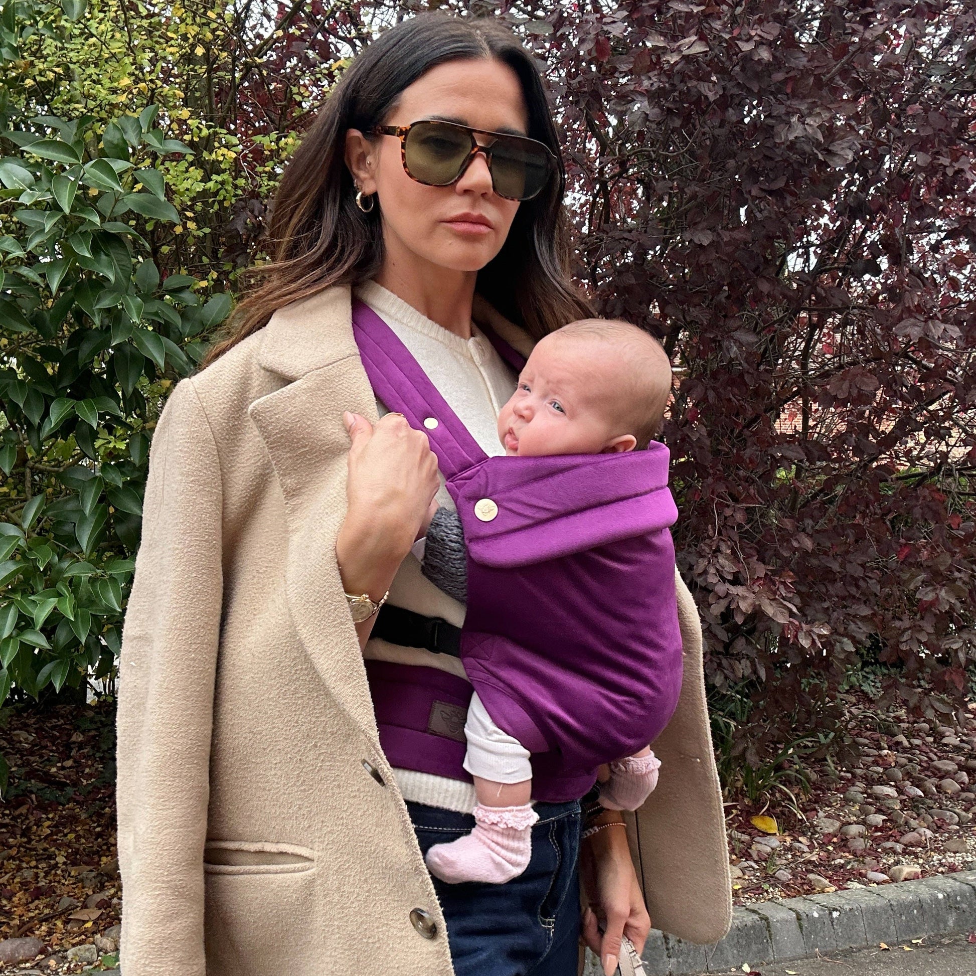 Woman holding a baby in a purple carrier outdoors.