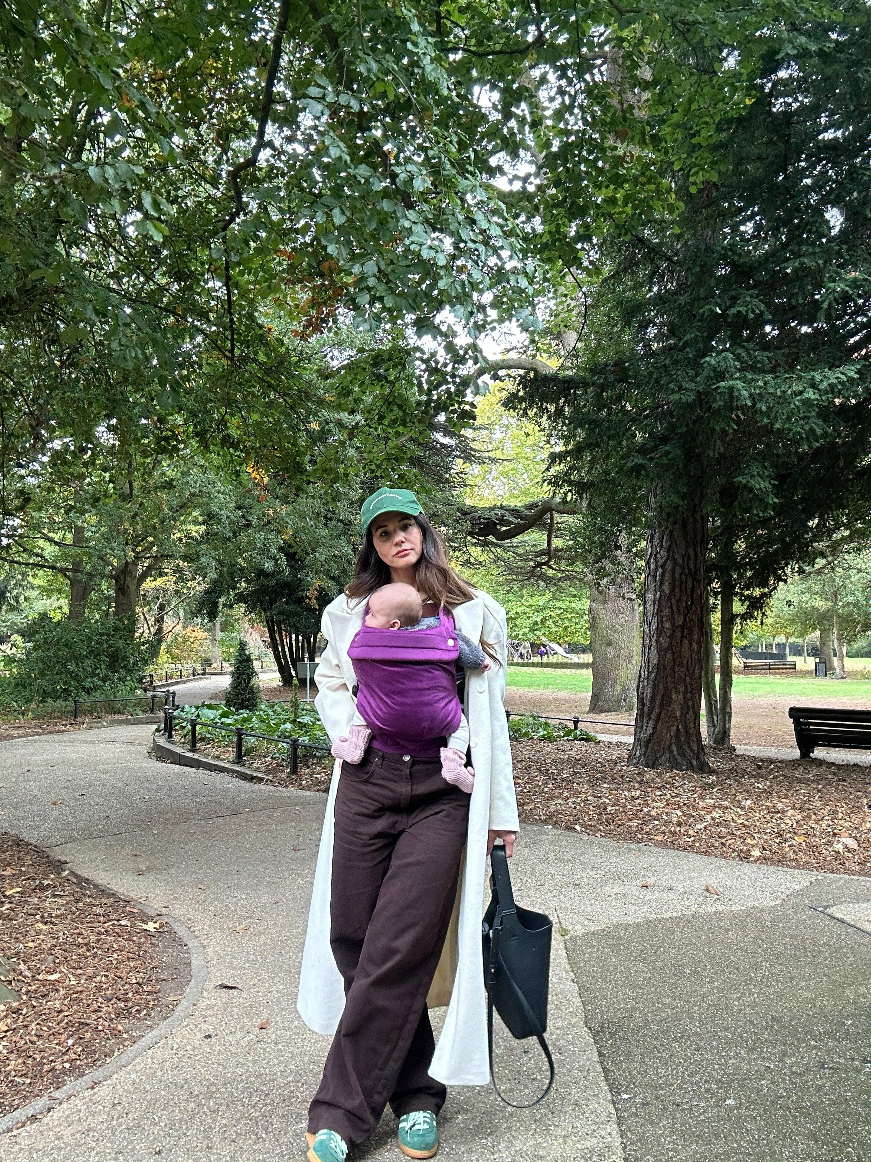 Woman walking in a park with a baby in a carrier, holding a bag.